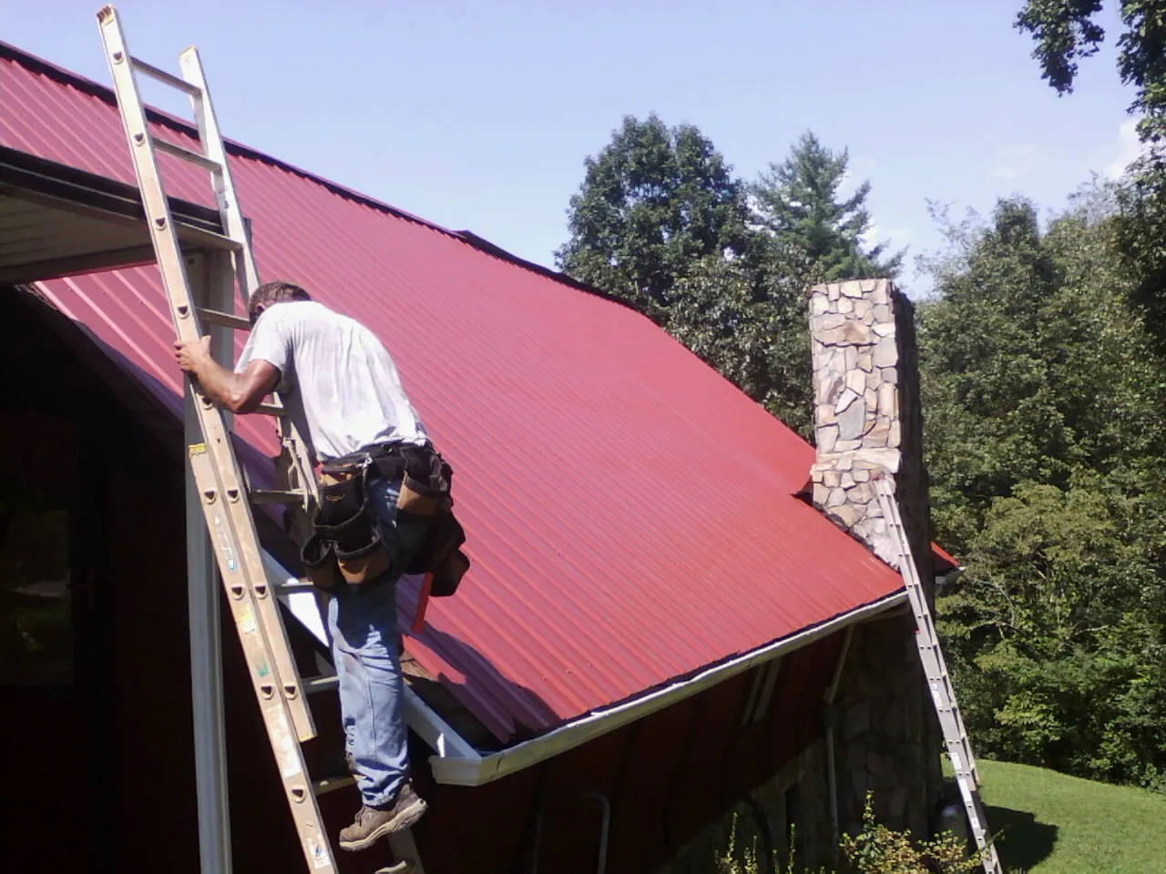 Roofer on a ladder beside a red metal roof and stone chimney — Soffit & Fascia Repair by Roofing Brothers in New City